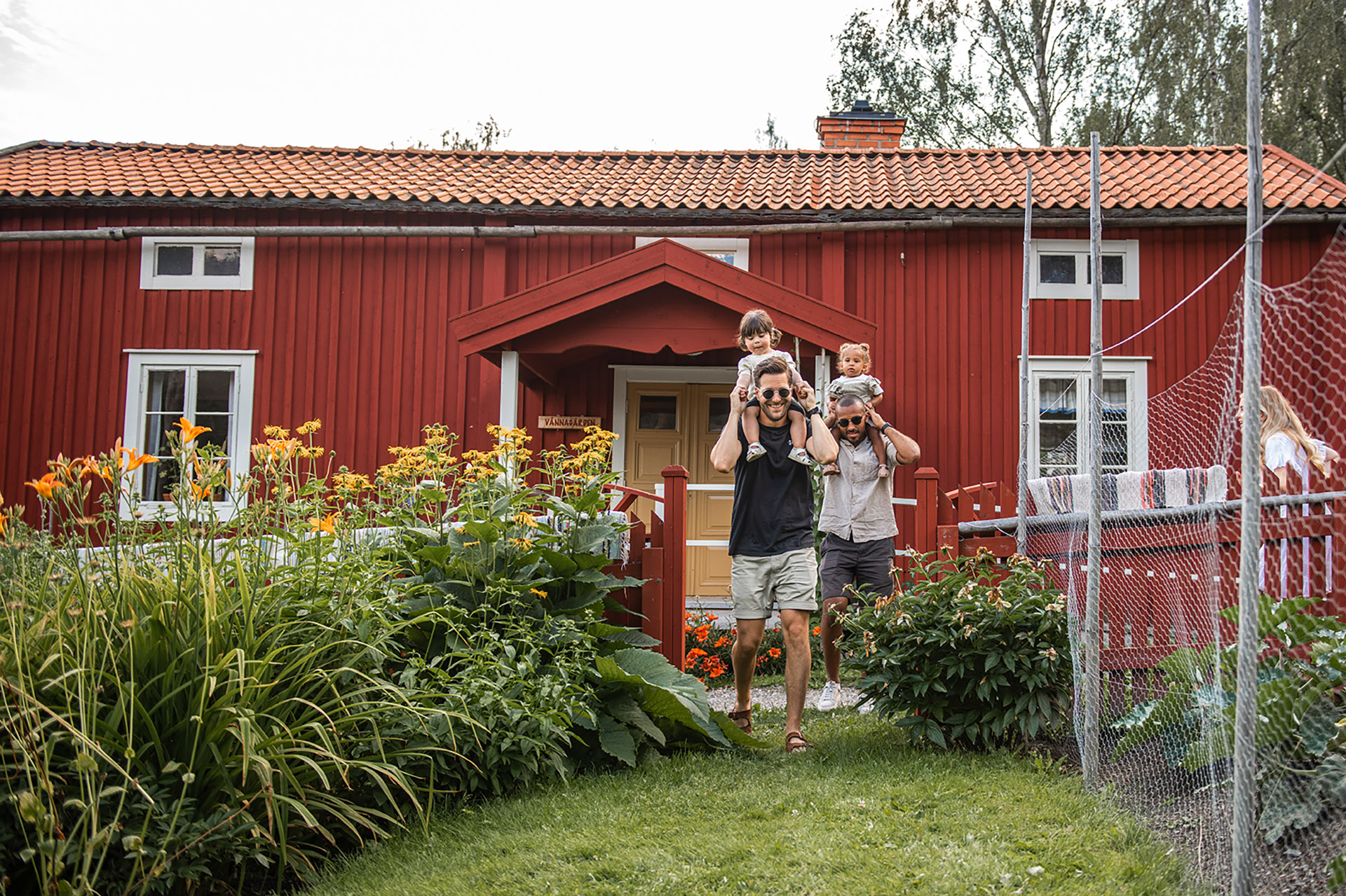 Två män med barn på axlarna går genom trädgård, med Vännagården i bakgrunden. Foto: Norra Berget.