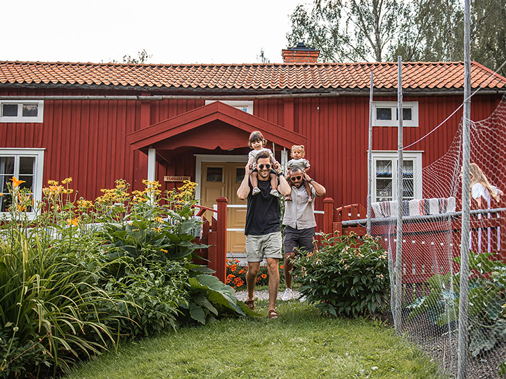 Två män med barn på axlarna går genom trädgård, med Vännagården i bakgrunden. Foto: Norra Berget.