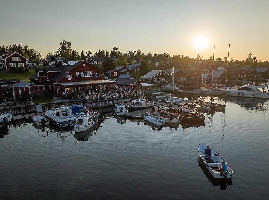 Fiskeläge med båtar intill brygga och uteservering med massor av människor.