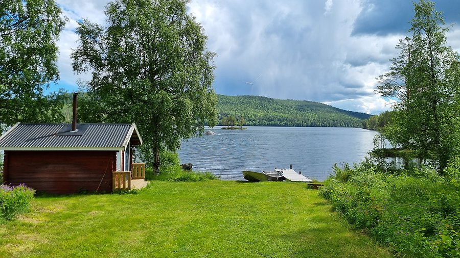 Röd liten stuga står intill en sjö med brygga.
