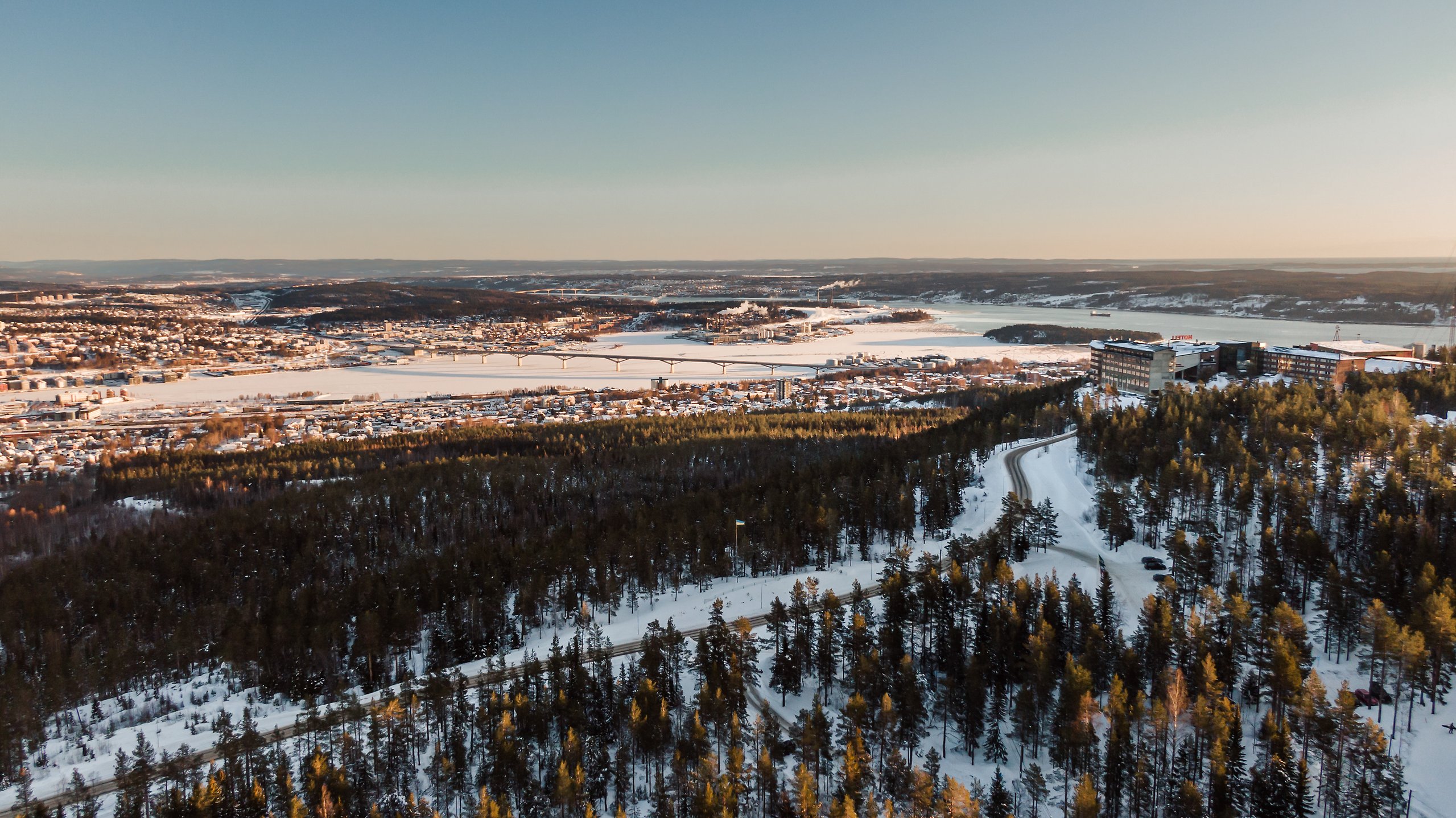 Ett foto från en drönare från toppen av Södra Berget. Snön ligger vit på marken, himlen är blå och solen skiner över Sundsvalls stenstad och hamn.