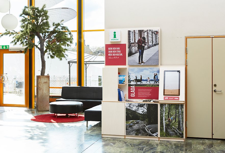 Foto av en entré. Det är stengolv och stora glaspartier. Till vänster i bild är ett träd i kruka och till höger står det en informationsdisk med digitalskärm. 