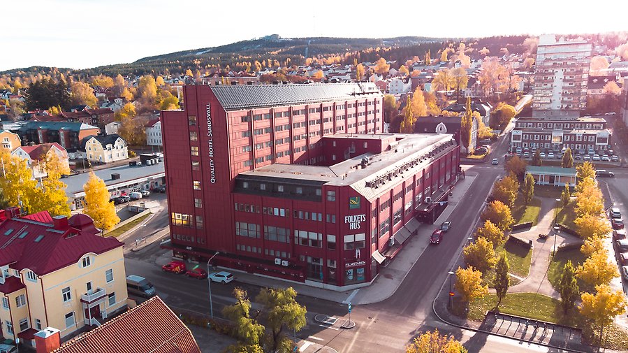 Fotografi taget av en drönare i luften. Fotografiet visar byggnaden Quality Hotell ligger i. man ser gator på sidan av huset samt en allé med träd.