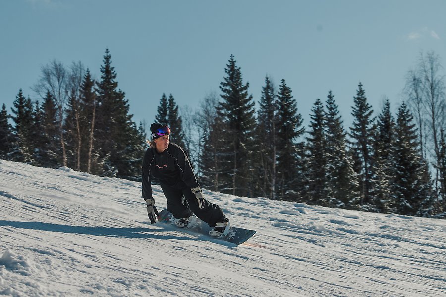 En man åker snowboard i en brant slalombacke. Pisten är snöigt vit och mannen är iklädd svarta kläder och har ett par stora glasögon på huvudet.