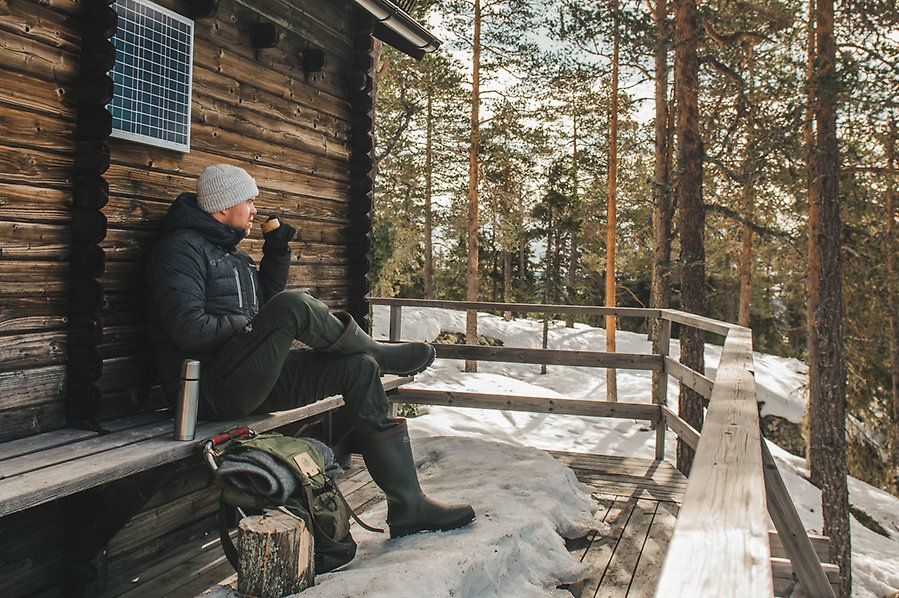 Fotografi av en person som sitter och dricker ur en mugg sittandes på en bänk utanför en stuga. Det är vinter och mycket snö.