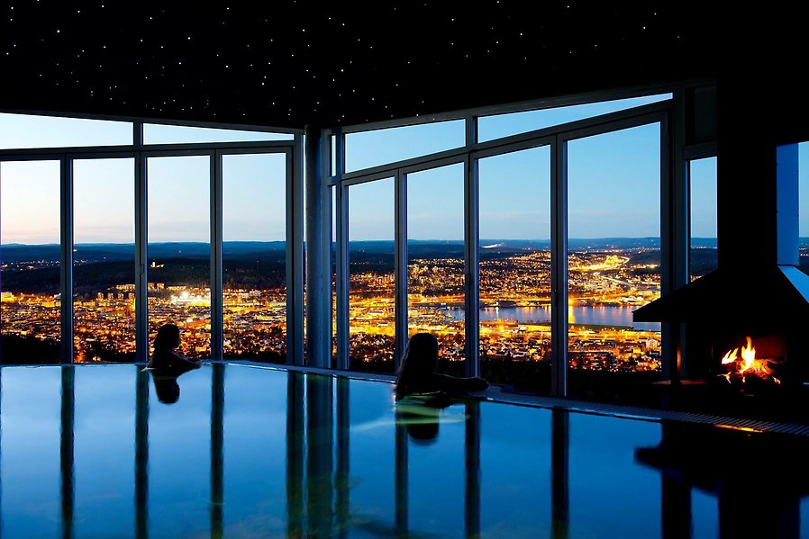 Foto av en inomhuspool med fönster bakom så att man ser ut över Sundsvalls stad. I poolen är det två personer som kolar ut på utsikten bakom poolen.